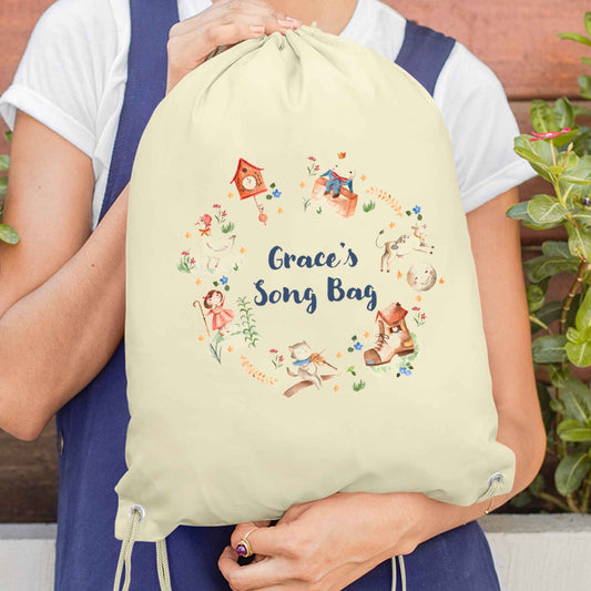 Image of woman holding a cream coloured cotton drawstring bag. Printed on the bag is some colourful illustrations from nursery rhymes, humpty dumpty, little bo peep, a clock, a boot, a goos, a cat and the fiddle and a cow and a moon. The watercolour illustrations are arranged in a wreath shape with the words 'Grace's son bang' written in blue in the middle.
