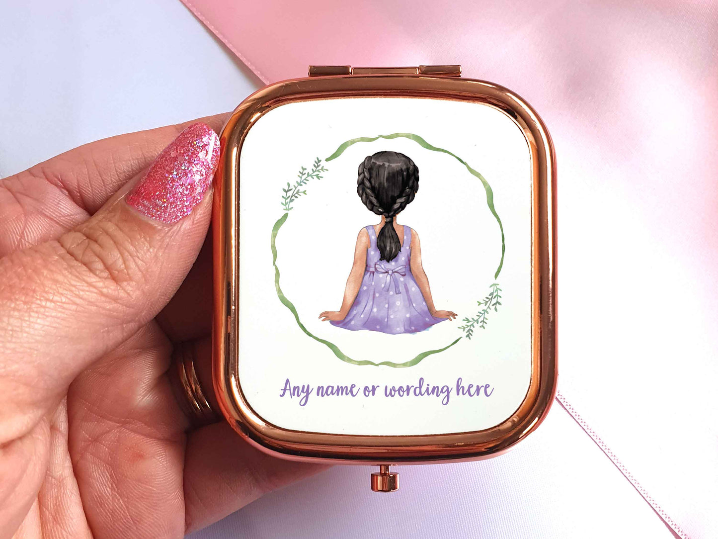 A hand holding a rose gold pocket mirror in a rectangular shape. A watercolour illustration of the back of a little girl is on the front of the mirror. The girl has black hair and tanned skin and is wearing a purple dress. Theres a green wreath around her, underneath in purple text it says 'any name or wording here'