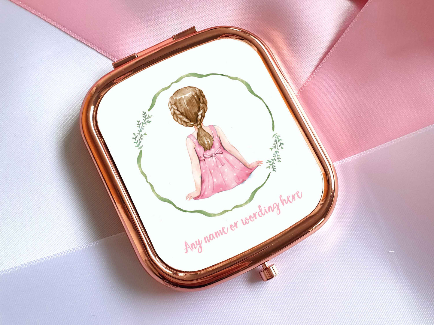 A rose gold pocket mirror in a rectangular shape. A watercolour illustration of the back of a little girl is on the front of the mirror. The girl has dark blonde hair and fair skin and is wearing a pink white polka dot dress. Theres a green wreath around her, underneath in pale pink text it says 'any name or wording here'