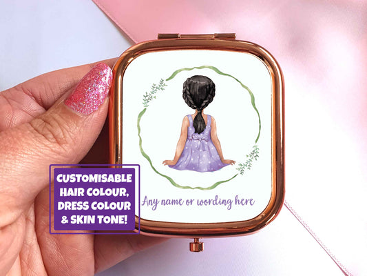 A hand holding a rose gold pocket mirror in a rectangular shape. A watercolour illustration of the back of a little girl is on the front of the mirror. The girl has black hair and tanned skin and is wearing a purple white polka dot dress. Theres a green wreath around her, underneath in purple text it says 'any name or wording here' 
On the image is a purple text box that says 'customisable hair colour, dress colour and skin tone'